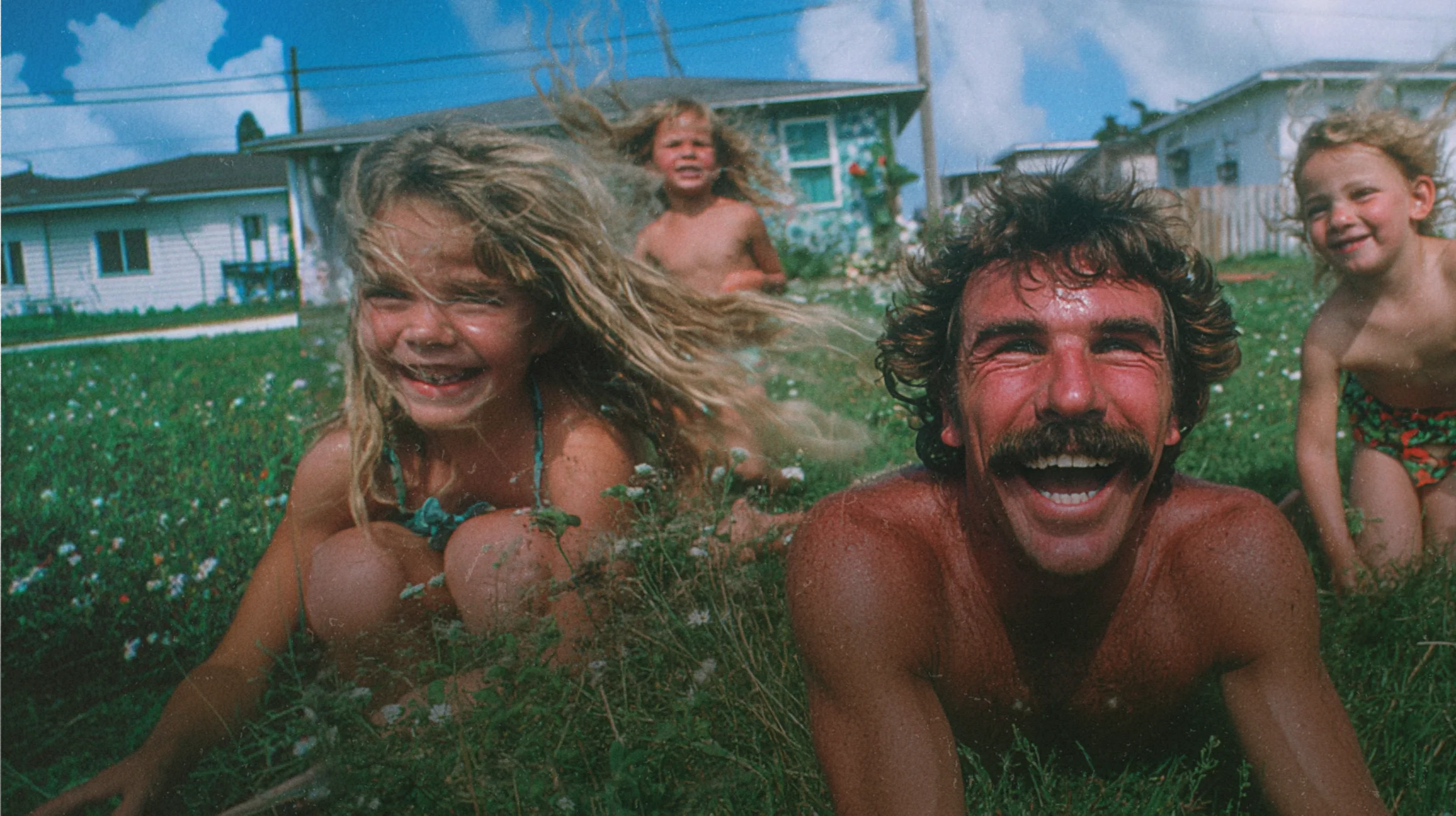 A dad and his two kids piled together on a green lawn, all laughing.