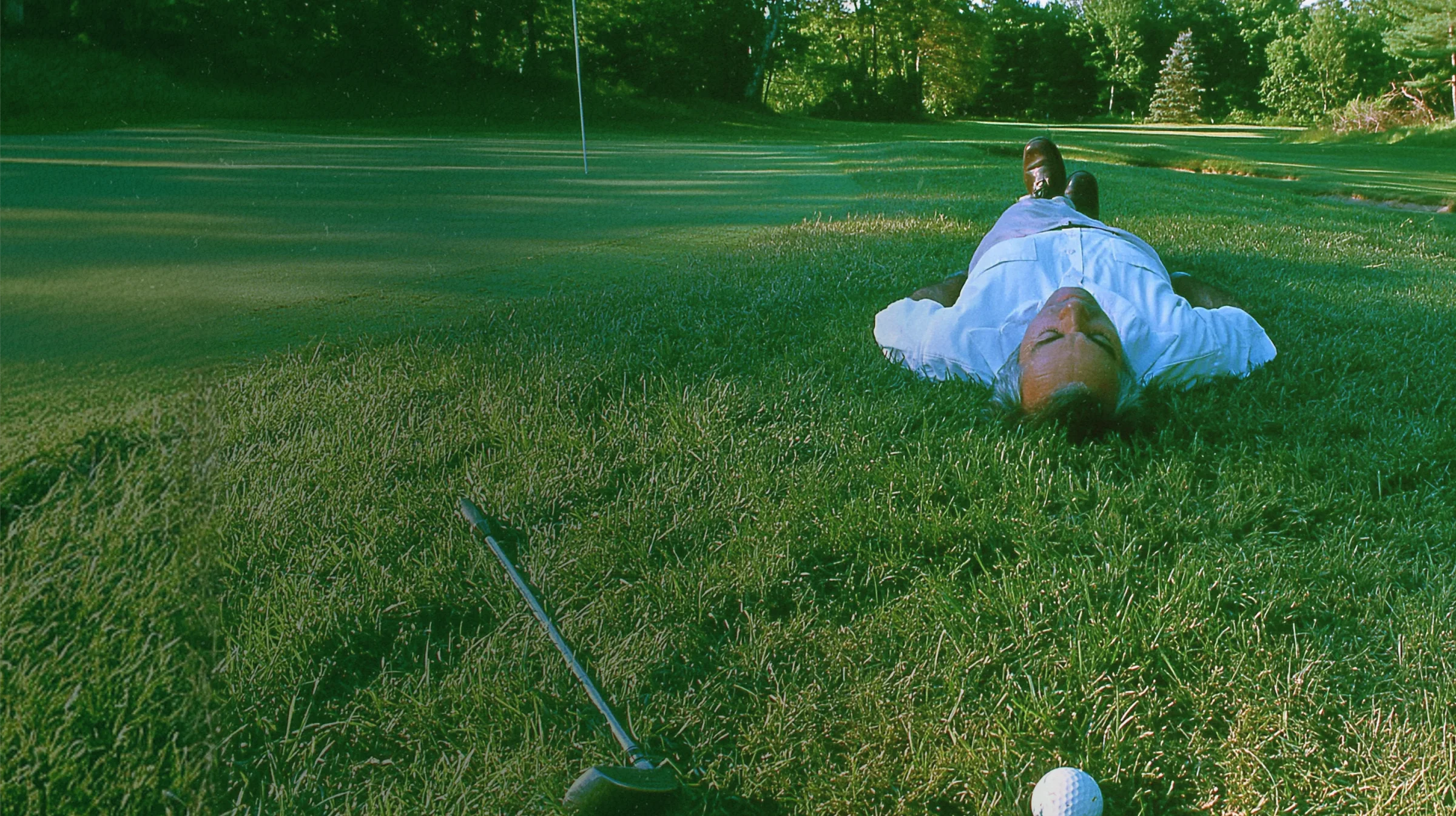 A man in formal attire napping on a putting green with a club and ball next to him.