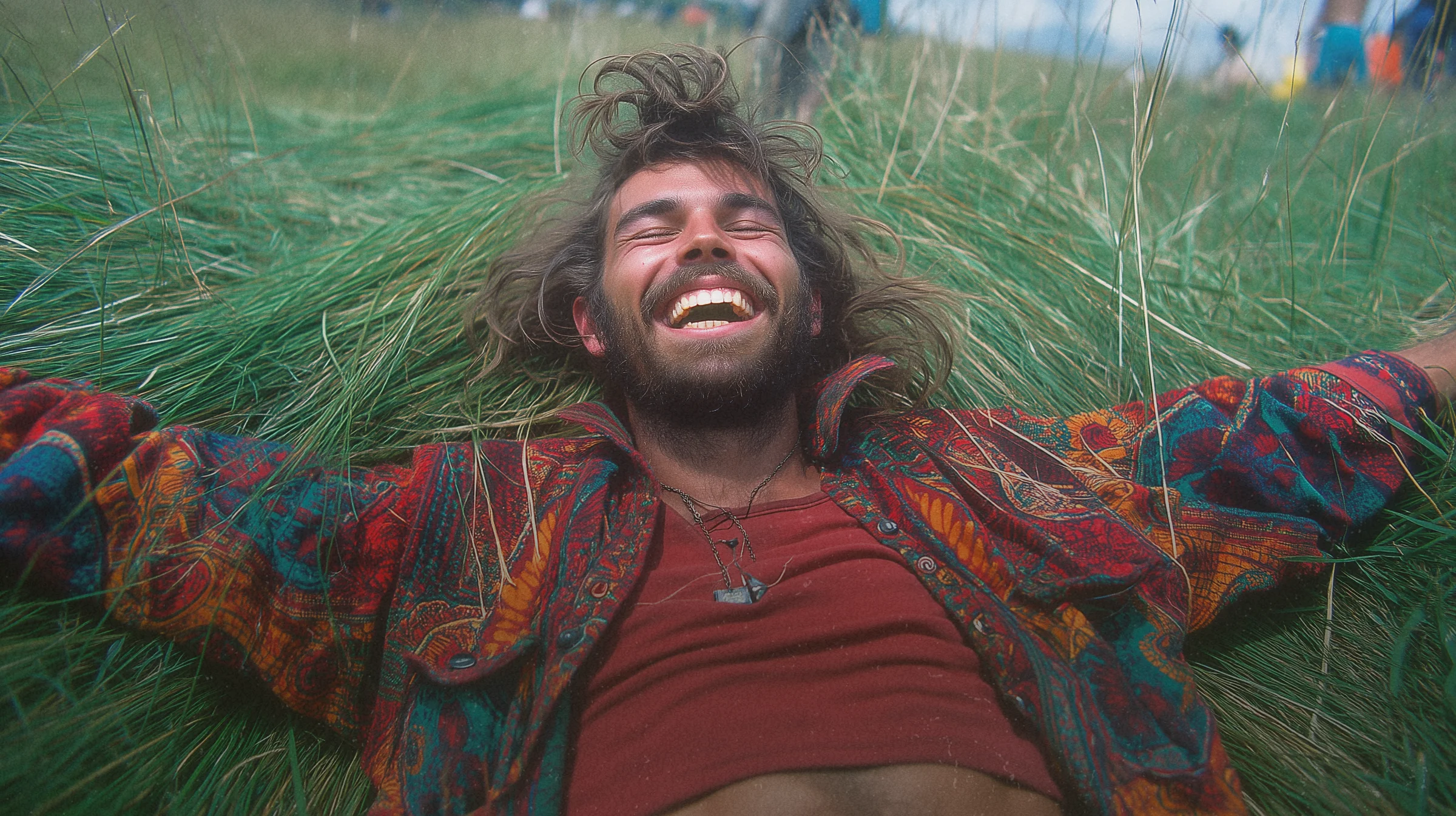 A long-haired man face-up in a thick green lawn, arms wide, blissed-out grin.