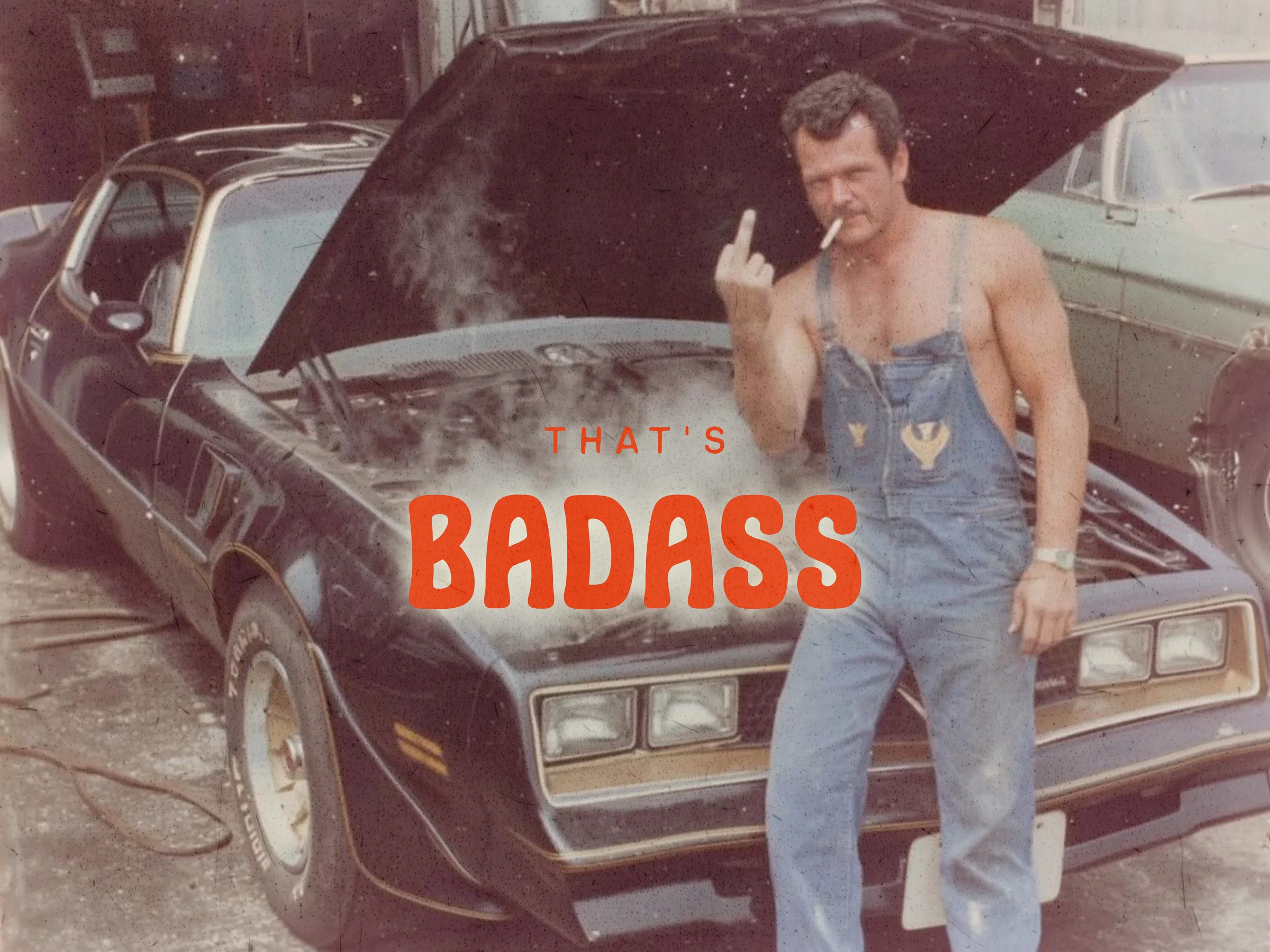 A shirtless mechanic in denim overalls smoking a cigarette and flipping off the camera next to a Trans Am, with the words THAT'S BADASS overlaid.